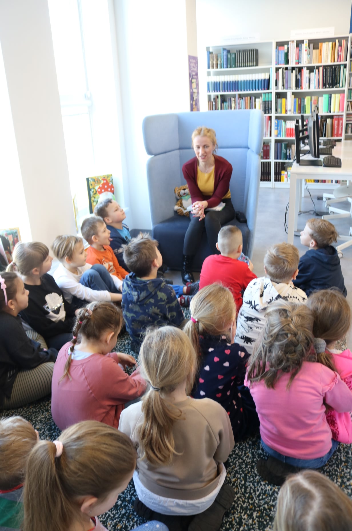 Dzieci siedzące na dywanie, słuchające bajke czytaną przez bibliotekarkę